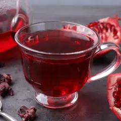 Aromatyczna herbata granat w szklance, obok świeże owoce granatu i suszone kwiaty hibiskusa.