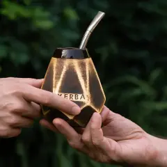 Dwie dłonie przekazują sobie ozdobną tykwę z napisem "YERBA" i metalową bombillą, gotowe do rytuału picia mate.