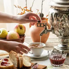 Dłoń odkręca kurek samowara, nalewając gorącą wodę do filiżanki. Obok jabłka, chleb z dżemem i dzban.