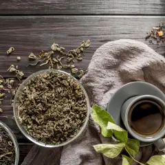 Na stole leżą liście herbaty, pokazując, gdzie rośnie herbata. Obok filiżanka z naparem i sitko.
