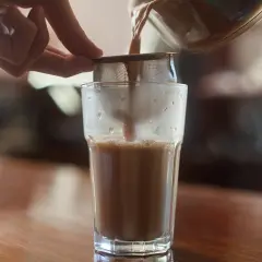 Ręka trzyma sitko, przez które przelewany jest aromatyczny masala czaj do szklanki.