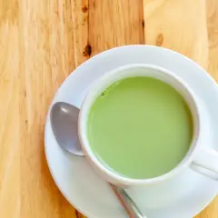 A creamy, vibrant zielona herbata z mlekiem w białej filiżance na drewnianym blacie. Obok leży łyżeczka.