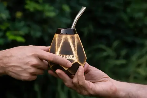 Dwie dłonie przekazują sobie ozdobną tykwę z napisem "YERBA" i metalową bombillą, gotowe do rytuału picia mate.