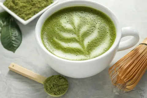 Zielona matcha w filiżance z pianką, obok miarka z proszkiem i bambusowa miotełka. Czy matcha ma kofeinę? Tak, zawiera ją!