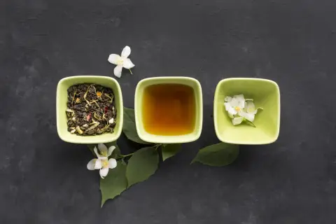 Trzy zielone miseczki na ciemnym tle: jedna z suszoną herbatą, druga z bursztynowym naparem, trzecia z białymi kwiatami jaśminu. Poczuj aromat koreańskiej herbaty.