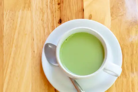 A creamy, vibrant zielona herbata z mlekiem w białej filiżance na drewnianym blacie. Obok leży łyżeczka.
