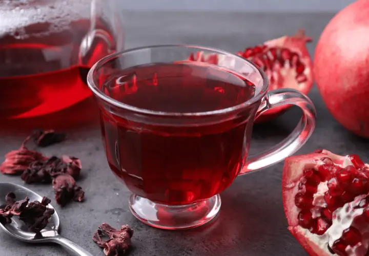 Aromatyczna herbata granat w szklance, obok świeże owoce granatu i suszone kwiaty hibiskusa.