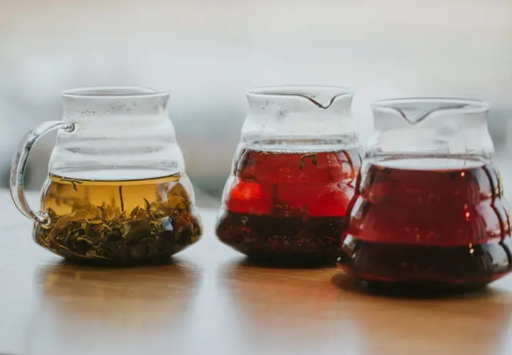 Trzy szklane dzbanki z zaparzonymi herbatami: jasnożółtą z liśćmi, czerwoną i ciemnoczerwoną, być może lapsang souchong.