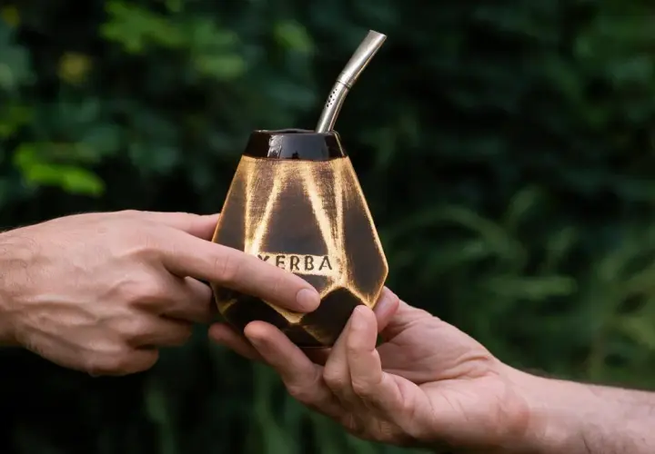 Dwie dłonie przekazują sobie ozdobną tykwę z napisem "YERBA" i metalową bombillą, gotowe do rytuału picia mate.