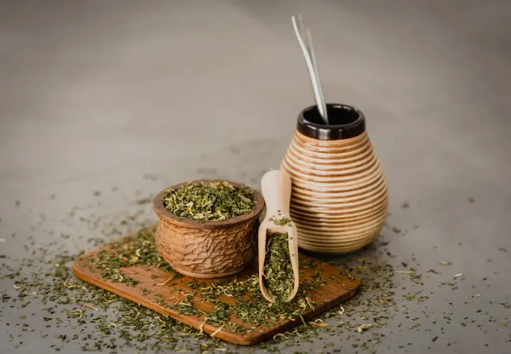 Drewniana miseczka i łopatka pełne suszonej yerba mate, obok ceramiczny naczynie z bombillą. Napar z yerba mate wspiera wątrobę.