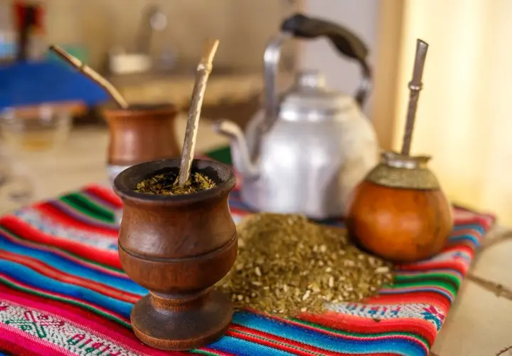 Tradycyjny rytuał z yaguar yerba mate: drewniane naczynia, bombille i czajnik na kolorowym obrusie.