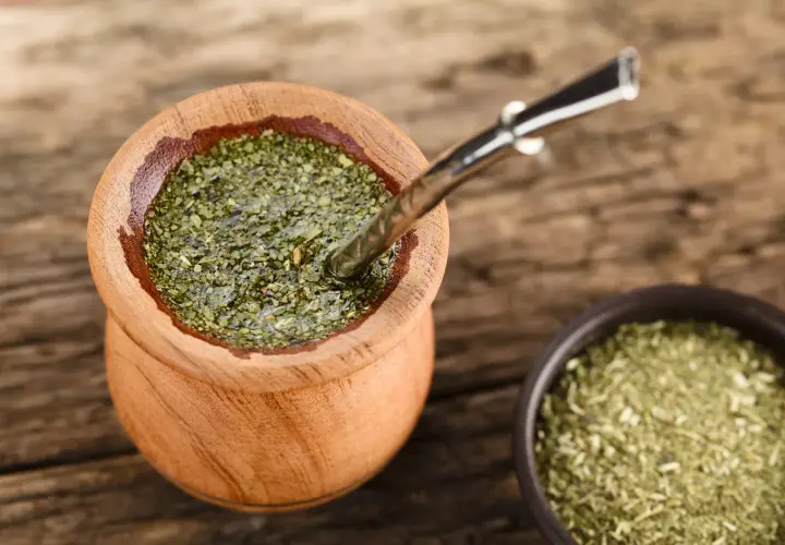 Drewniane naczynie z yerba mate na uspokojenie, obok miseczka z suszem. Metalowa bombilla zanurzona w naparze.