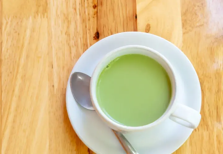 A creamy, vibrant zielona herbata z mlekiem w białej filiżance na drewnianym blacie. Obok leży łyżeczka.