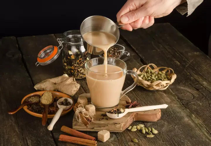 Ręka wlewa mleko do kubka, tworząc aromatyczną herbatę po irlandzku. Wokół przyprawy: cynamon, kardamon, imbir.