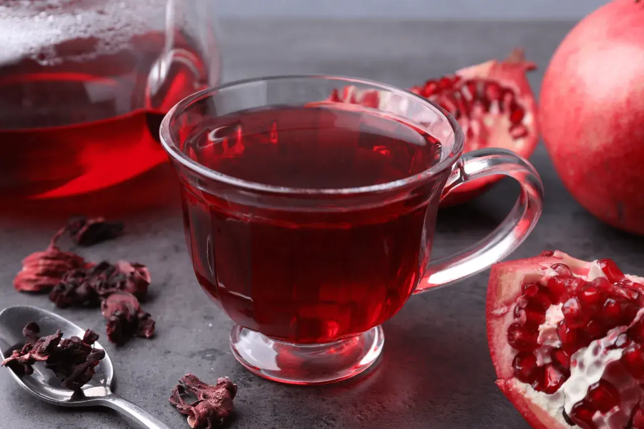 Aromatyczna herbata granat w szklance, obok świeże owoce granatu i suszone kwiaty hibiskusa.