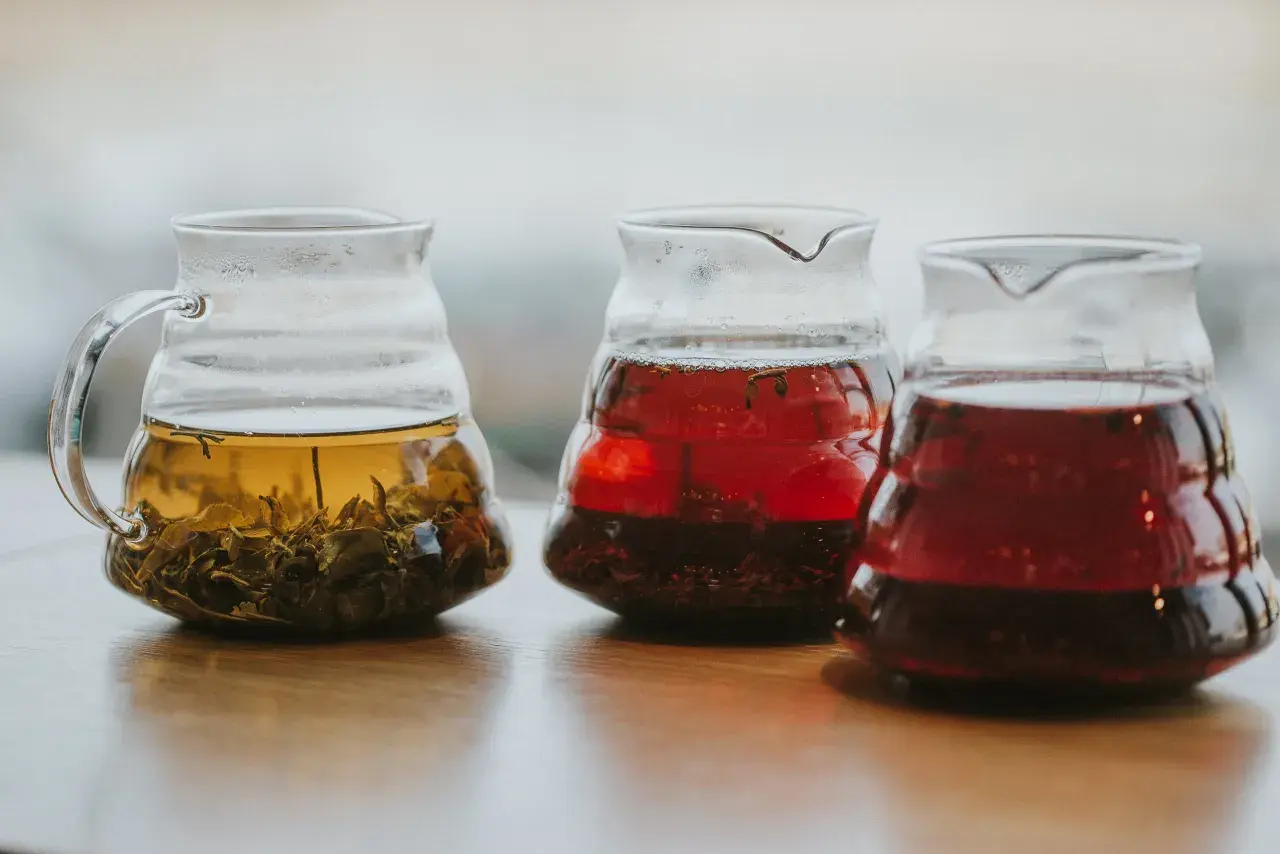 Trzy szklane dzbanki z zaparzonymi herbatami: jasnożółtą z liśćmi, czerwoną i ciemnoczerwoną, być może lapsang souchong.