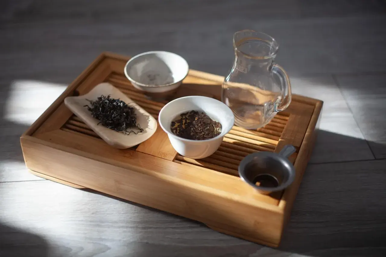 Zestaw do gongfu cha: drewniana taca, liście herbaty, czajniczek, miseczki i sitko.