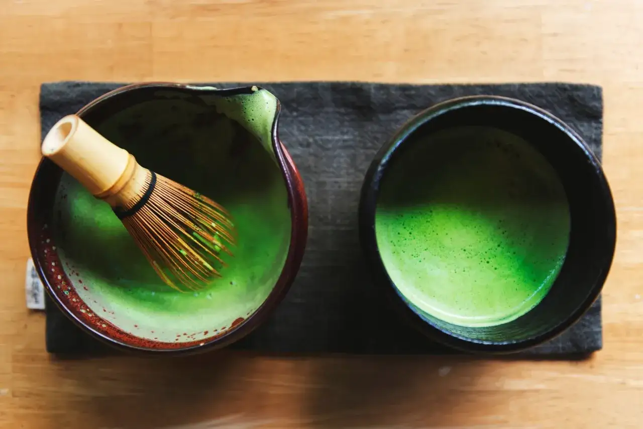Przygotowanie idealnego matcha: bambusowa miotełka w miseczce z zielonym proszkiem i wodą, obok gotowy napój.