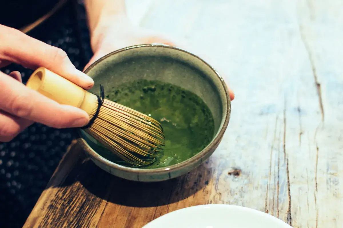 Ręka miesza bambusową miotełką zieloną matchę w miseczce. Tak przygotowana matcha ma intensywny, lekko gorzkawy smak.