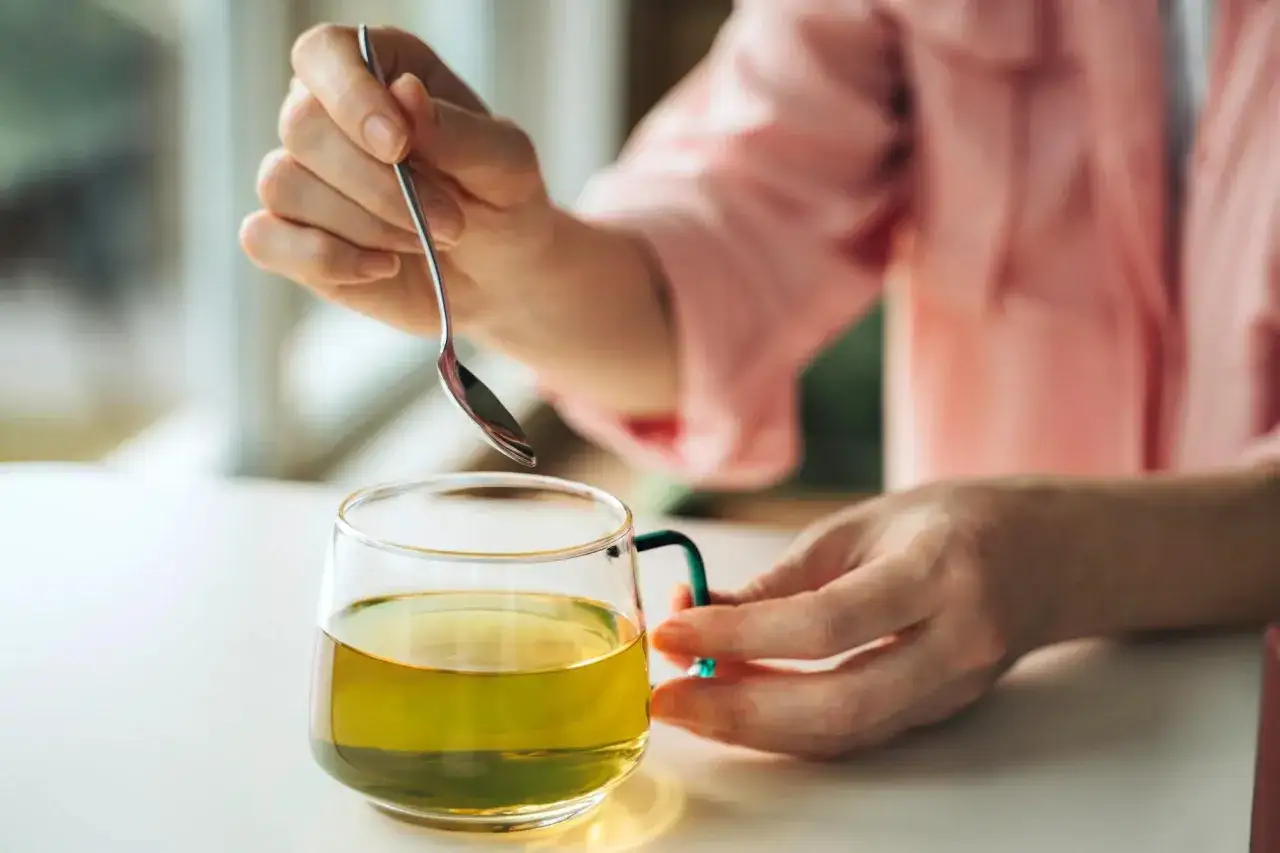 Ręka miesza łyżeczką aromatyczną zieloną herbatę na noc w szklanym kubku.