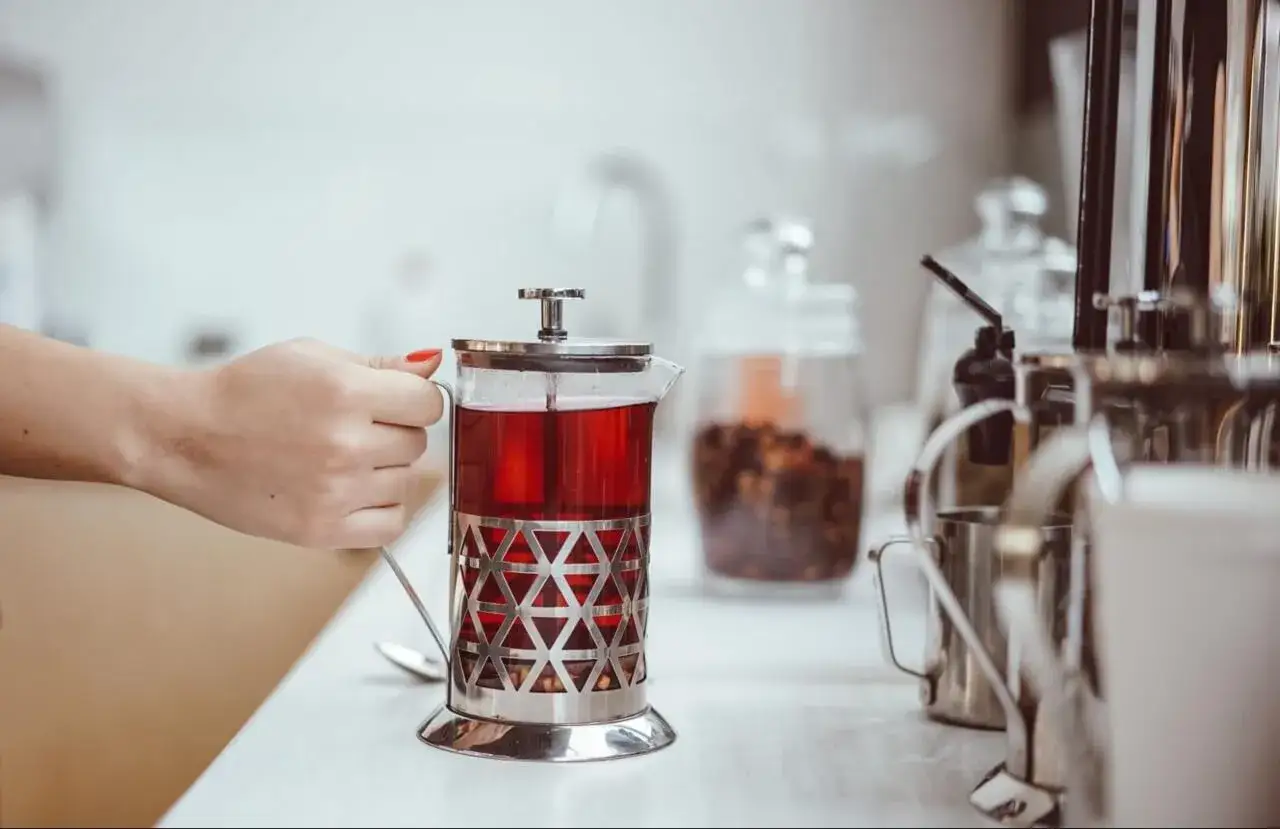 Dłoń trzyma szklany french press z aromatyczną, czerwoną herbatą.