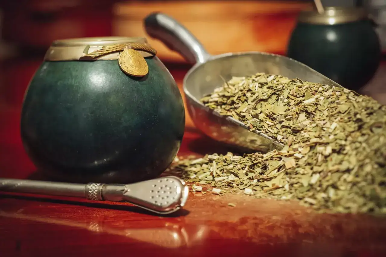 Zielona tykwa z ozdobnym medalionem, obok metalowa łopatka z suszem yerba mate i bombilla.