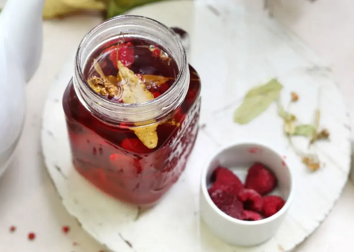 Orzeźwiająca domowa ice tea z malinami i lipą w słoiku, obok miseczka z mrożonymi malinami. Idealna na upalne dni.