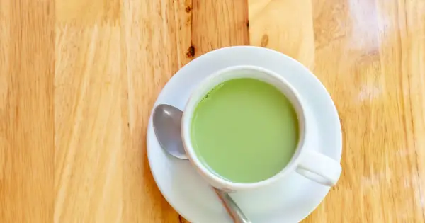 A creamy, vibrant zielona herbata z mlekiem w białej filiżance na drewnianym blacie. Obok leży łyżeczka.